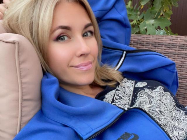 Misstyk with blonde hair, blue eyes, and pink lips, wearing a blue jacket with black and white pattern, smiling slightly, resting on a beige pillow.