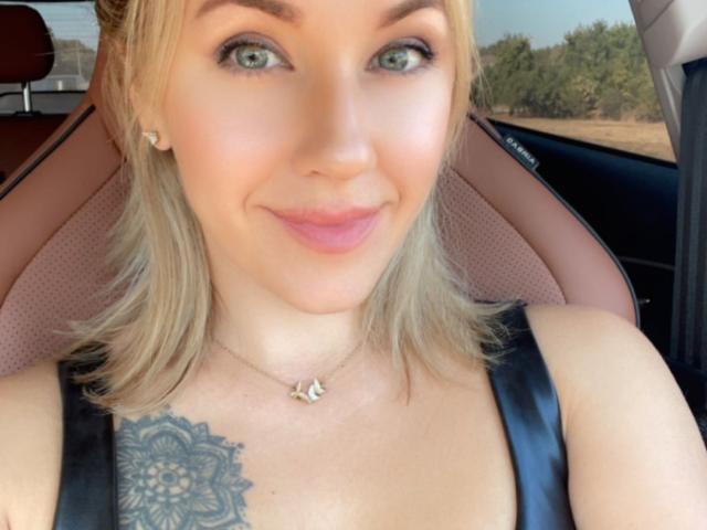 Misstyk with blonde hair, green eyes, and a subtle smile, wearing a black leather top, showing a detailed tattoo on her right boob, sitting in a car.