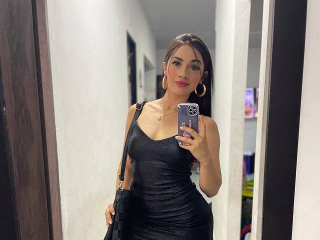 AngelCutie avec une poitrine de taille moyenne, portant une robe noire moulante, tenant un téléphone, avec du rouge à lèvres, des boucles d'oreilles cerceaux et un sac bandoulière noir, prenant un selfie devant un miroir..