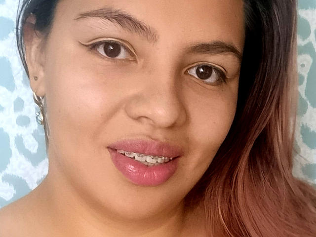 YinethBeilly has medium brown skin, dark brown eyes, and pink glossy lips with visible teeth. She has long brown hair, a silver earring, and subtle winged eyeliner.