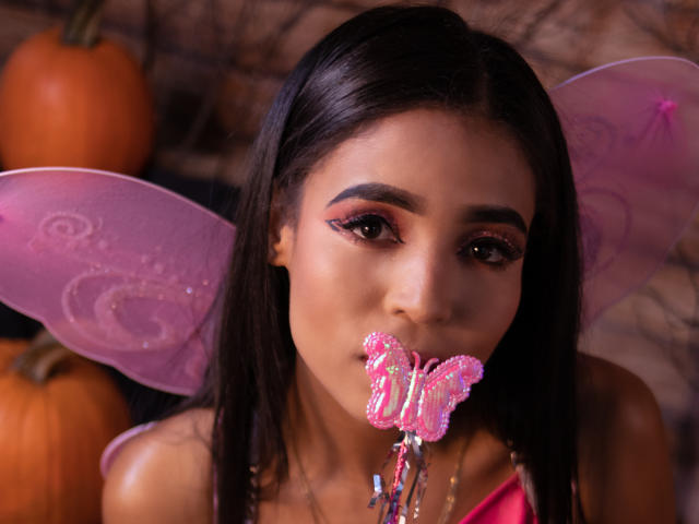 LaylaBrowns with dark hair, pink fairy wings, and pink butterfly-shaped candy in her mouth, holding colorful strings. Her makeup is bold with pink eyeliner.