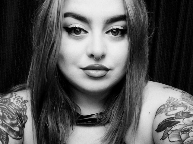 ChaudePourxToi with long, slightly messy hair, thick eyebrows, winged eyeliner, nose ring, and pouty lips, showing tattoos on her shoulders. She's wearing a black choker.