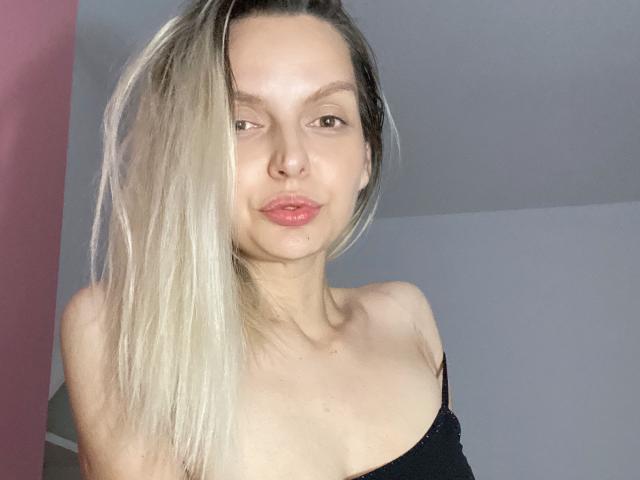 DouceAmelia with blonde ponytail, fair skin, small nose, pink lips, wearing black strap top, visible collarbones, light in face, slight smile, looking at camera.