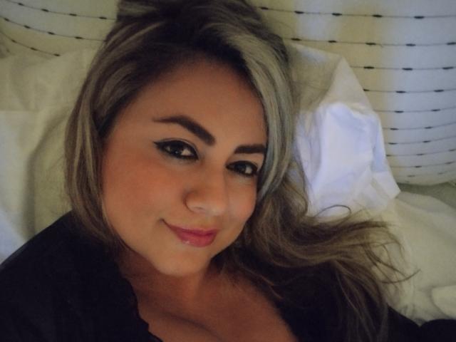 AmandaLeon with blonde hair, black eyeliner, and pink lipstick, smiling at the camera, lying in bed, showing her cleavage in a black top.