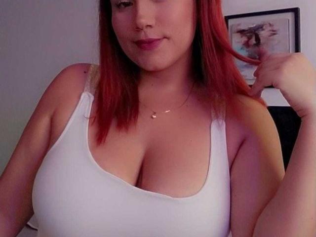 SharonGreys mit roten Haaren, die ein weißes Tanktop trägt und ihre großen Brüste zeigt, lächelt leicht, hält eine Haarsträhne und trägt eine kleine Halskette, in einem schwach beleuchteten Raum..