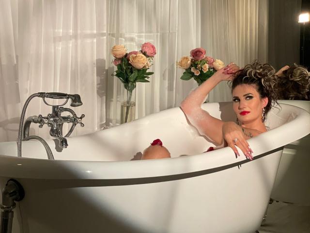 Evelyn Frances in een wit bad, met rozenblaadjes, rode lippenstift en krullend haar, die een arm omhoog steekt, waarbij haar borsten zichtbaar zijn, met een zilveren kraan en bloemen op de achtergrond..