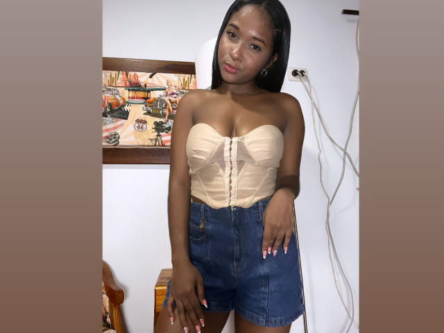 MelanyScoth ha la pelle scura, lunghi capelli neri e indossa un corpetto beige senza spalline con bottoni e pantaloncini di jeans a vita alta, in piedi con le mani vicino all'inguine..