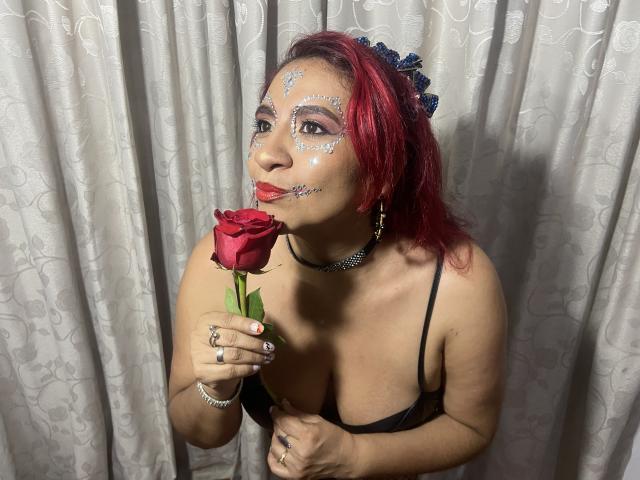 NahelaBigMelonsX with red hair, glitter face, black lingerie, holding red rose to mouth, wearing blue hair clip and choker, standing against white curtains, showing medium tits.