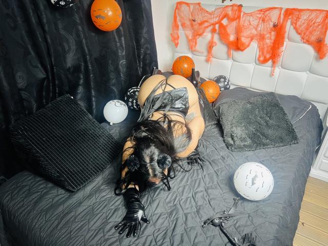 CarlaSexyFontaine su un letto, vestita con lattice nero, guanti e una maschera, a faccia in giù, con il sedere rivolto verso l'alto, con palloncini arancioni e tessuto arancione sullo sfondo..