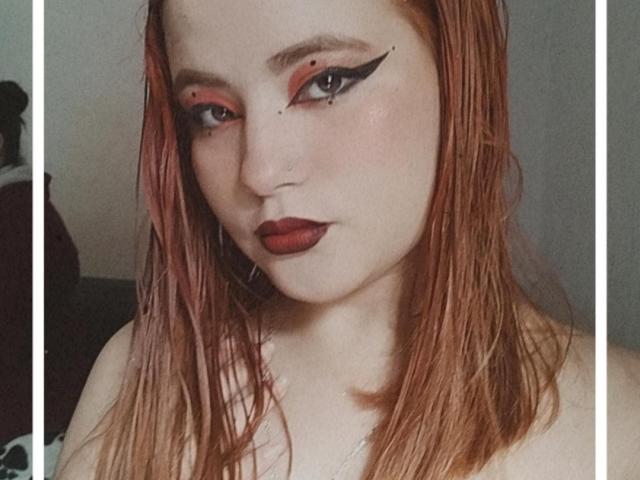 VenusCrazzy ha i capelli rossi, l'eyeliner nero applicato sulle palpebre e il rossetto rosso. Sta guardando la fotocamera con un'espressione seria, mostrando il piercing al naso. Il suo seno non è visibile..