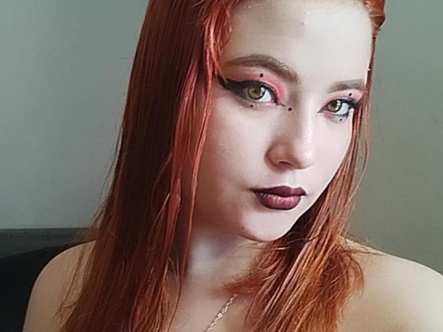 VenusCrazzy ha i capelli rossi, gli occhi verdi con l'eyeliner nero e il rossetto viola scuro. Sta guardando la fotocamera con un sorriso leggermente seducente e sicuro di sé..