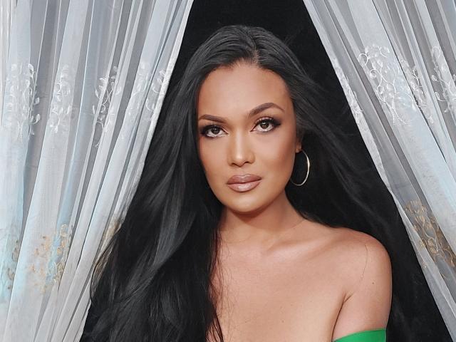 DreamOfDarkness has long black hair, brown eyes, and is wearing a green off-shoulder top with hoop earrings, standing between sheer curtains. Boobs are partially visible.