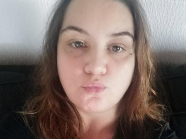 Sylvia69 tiene la piel clara, ojos marrones y cabello ondulado de color marrón, con una pequeña peca sobre su ojo derecho. Está frunciendo los labios y lleva una blusa negra..