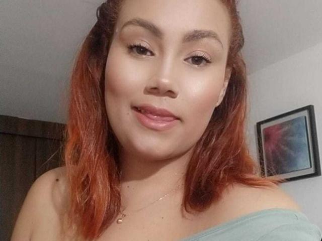 SharonGreys hat rot-orangefarbenes Haar, helle Haut und lächelt mit pinkfarbener Lippenstift. Sie trägt ein grünes Oberteil mit einem Schulterauschnitt, das ihre nackten Schultern zeigt, und eine kleine Halskette..