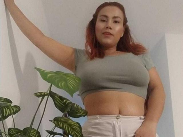 Sharon Grey mit roten Haaren, einem grünen Crop-Top, weißen Hosen und einem sichtbaren Bauch, steht mit einem Arm erhoben neben einer grünen Pflanze mit gestreiften Blättern..