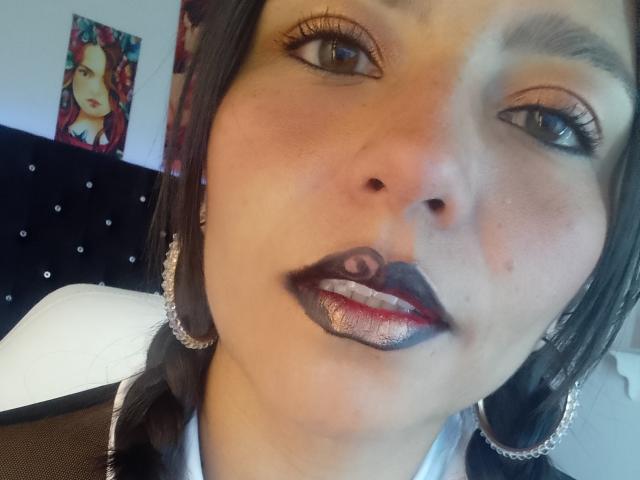 El rostro de Becck, con un labial oscuro con brillo, detalles en rojo y negro, y aros grandes, con cabello negro trenzado y pestañas largas, en una foto tipo selfie..