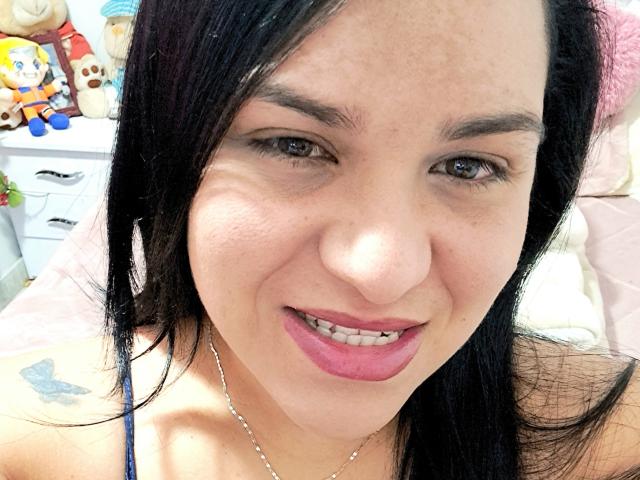 VivianaThompson has black hair, pink lipstick, and a smile showing her teeth. She's wearing a blue spaghetti strap top and a silver necklace with a butterfly tattoo.