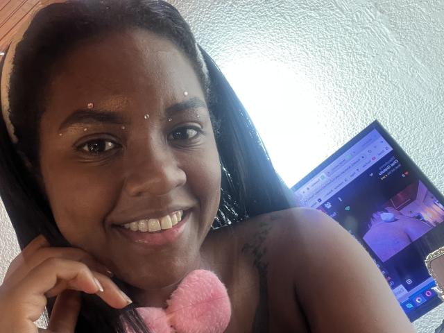 TiffanySammy with dark skin, smiling, wearing a pink heart-shaped necklace, sparkling facial dots, black hair, holding phone, tattoo on shoulder, TV screen in background showing video.
