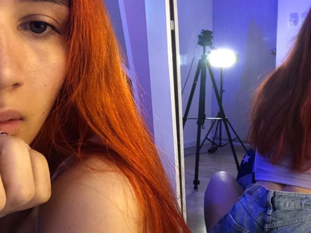 Greta Harris, com cabelos ruivos longos, olhando para um espelho, mostrando seu reflexo, com uma luz de tripé ao fundo, vestindo shorts de jeans e mordendo o dedo..