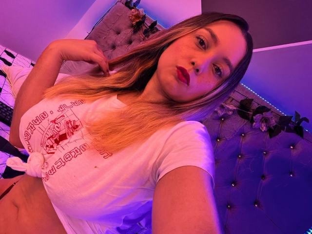AmayaJhons with blonde hair, red lipstick, white tied shirt, pointing at her chest, purple and pink lighting, showing her tits, in a bedroom with a tufted headboard.
