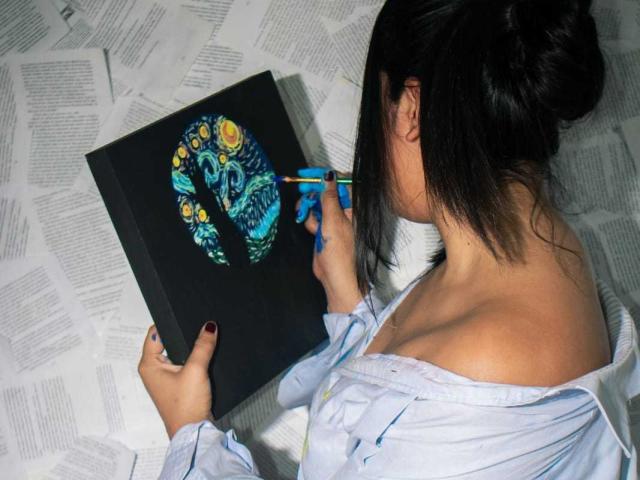 Rossy Granger, con i capelli scuri raccolti in un disordinato chignon, sta dipingendo un colorato ritratto del volto di una donna su una tela nera, usando un pennello blu, indossando una camicia bianca con una spalla scoperta..
