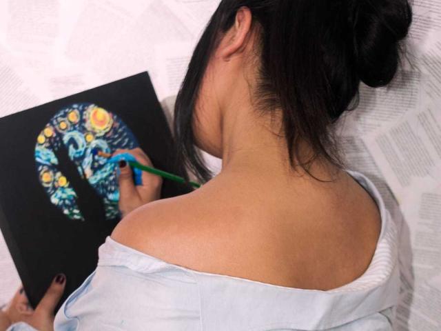 RossyGranger with black hair in a bun, bare shoulders, white shirt, painting with colorful, swirling patterns, holding green pencil, papers scattered behind.