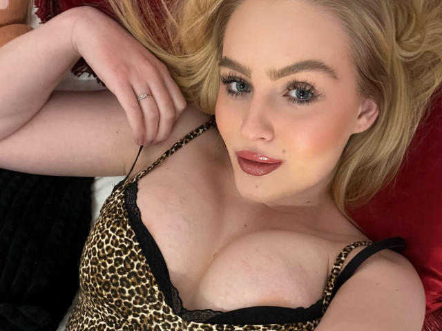 Olivia: Een vrouw met blond haar, blauwe ogen en glanzende, roze lippen, gekleed in een top met luipaardprint, die haar grote, glanzende borsten laat zien, liggend op een rode ondergrond..