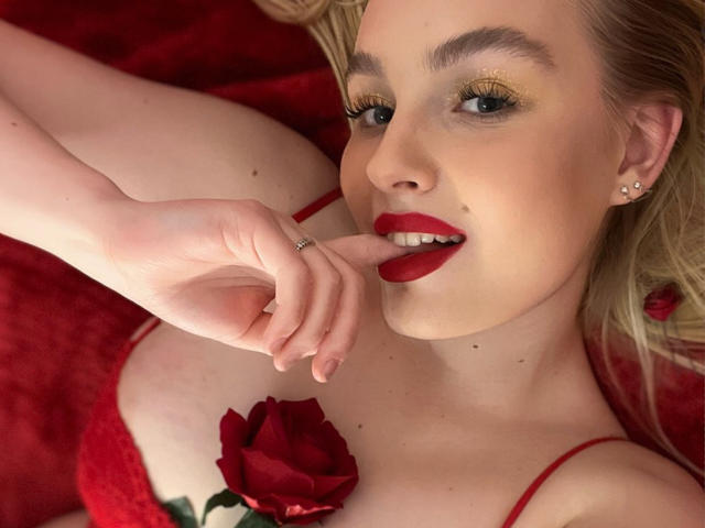 OliviaBent with blonde hair, red lipstick, and earrings, biting her finger, red rose near her tits, red lingerie, and golden eyeshadow, lying on red fabric.