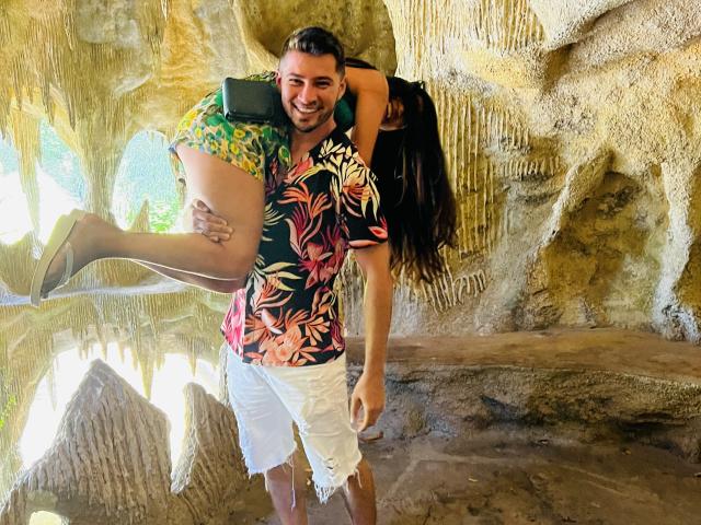 JuanAndLiza, gekleed in een bloemenhemd, draagt een vrouw met gele shorts en een groene top. Haar billen zijn omhoog, ze glimlacht en heeft bruin haar. Juan heeft zijn arm om haar middel. De setting lijkt op een grot..