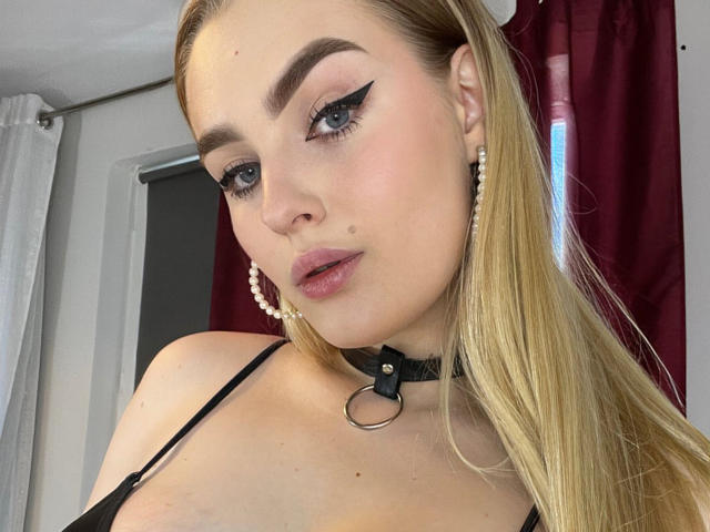 OliviaBent tiene el cabello rubio, delineador de ojos negro y aretes de perlas, lleva un collar negro con un anillo, y una blusa negra que muestra sus senos y una pequeña marca de nacimiento..
