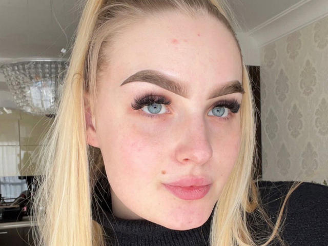 Olivia Bent tiene ojos azules, una coleta rubia, cejas pobladas, pestañas largas, lápiz labial rosa y una pequeña marca de nacimiento en su mejilla izquierda. Lleva un jersey de cuello alto negro..