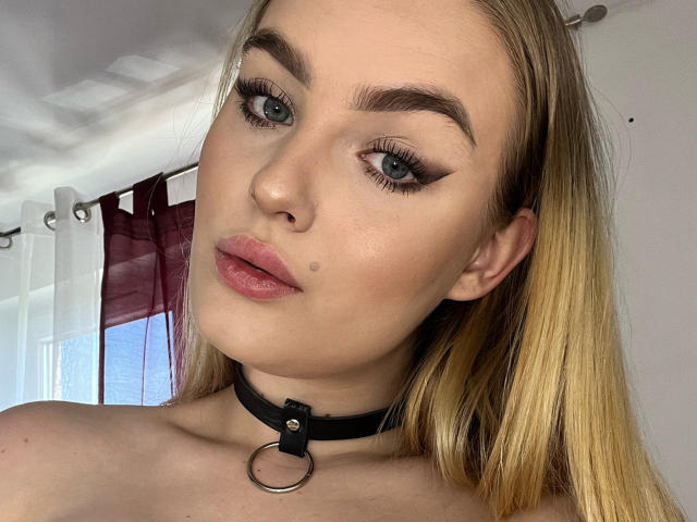 Olivia, avec ses cheveux blonds, ses yeux bleus et ses lèvres roses, porte un collier noir avec un anneau métallique, a une petite tache de beauté sur sa joue droite, et regarde l'appareil photo..