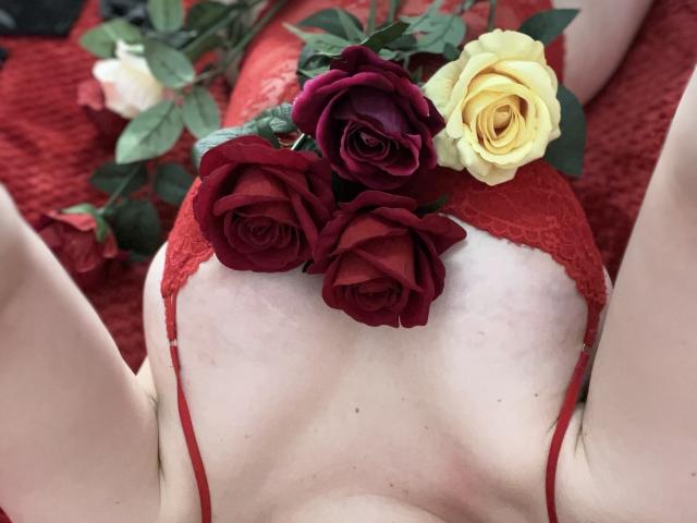 O sutiã de renda vermelha de OliviaBent, cobrindo seus seios, com três rosas — duas vermelhas e uma amarela — colocadas em seu peito. Lençol vermelho no fundo..