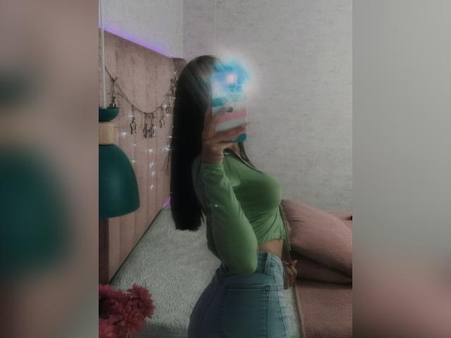 AnyaJay con lunghi capelli castani, una maglia verde a maniche lunghe, jeans blu a vita alta, con il telefono davanti che le copre il viso, seduta su un letto rosa, che mostra i suoi fianchi prosperosi e i piccoli seni..