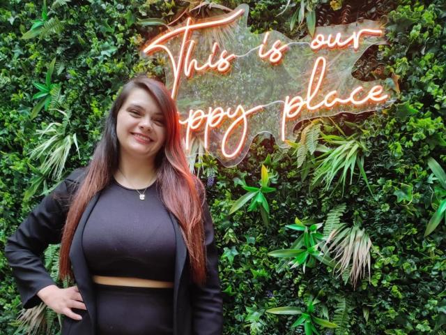 katherinLatin, with long brown hair, black crop top, and black jacket, stands smiling in front of neon sign, green wall of plants, and has hands on hips.