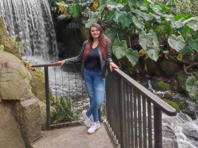 KatherinLatin stands by a waterfall, wearing a black jacket, blue jeans, and white sneakers, with long brown hair and a smile, her right hand on a metal railing.