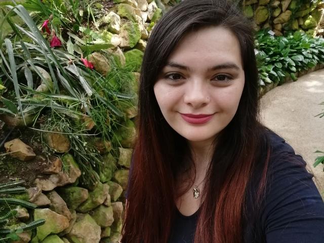 KatherinLatin with long brown hair, pink lips, and dark eyes, wearing a black shirt, smiling at the camera, has a necklace with a small pendant, and a cute nose.