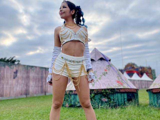 Megan Wisse, con piel morena, cabello negro recogido en una coleta, pechos pequeños, vestida con una camiseta blanca corta adornada con una cadena dorada, pantalones cortos y guantes blancos largos, de pie en un campo cubierto de hierba..