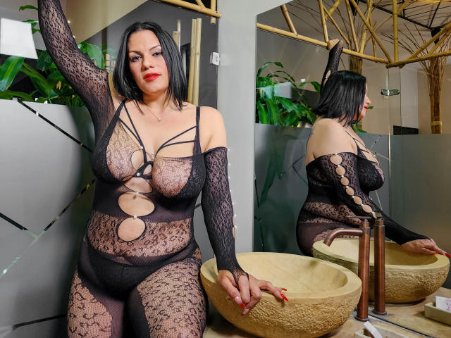 LizaRussell with black lace bodysuit, showing big tits and round ass, standing by a bathroom sink, her black bob and red lipstick contrasting with her pale skin.