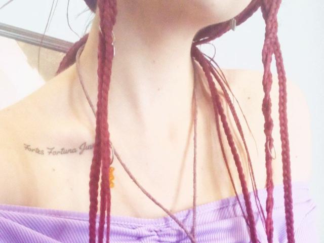 Le trecce rosse di Zanoza le scendono lungo i fianchi, indossa una maglietta viola con una spalla scoperta, mostrando un tatuaggio appena sotto il seno, con una collana e perline arancioni..
