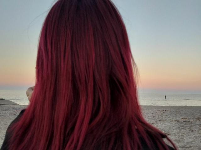 Zanoza's red, straight, medium-length hair covers her back, facing a sunset over a pebble beach with a small person in the distance.