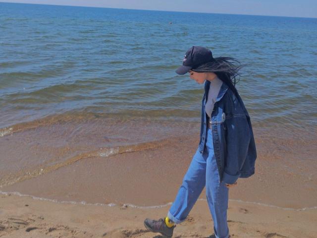 Zanoza walking on a sandy beach, wearing a black cap, blue jacket, and jeans, with a black backpack, and gray shoes, with her head turned to the side.
