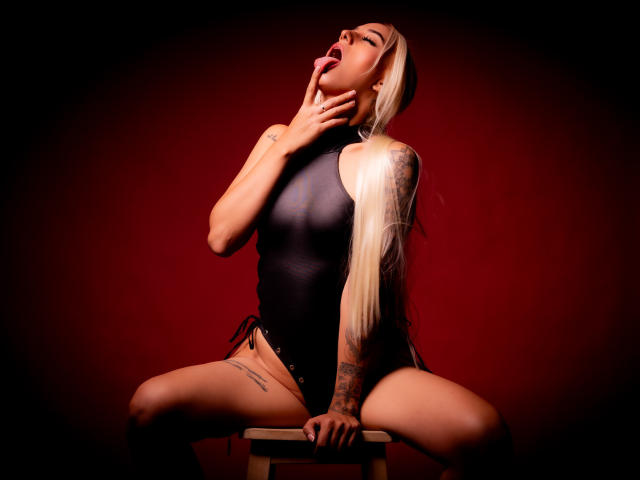 AnnieLaurenn, avec de longs cheveux blonds, la langue tirée, une main sur le cou, un haut noir sans manches, un pantalon noir à lacets, un bras tatoué, assise sur un tabouret, fond rouge, jambes écartées..