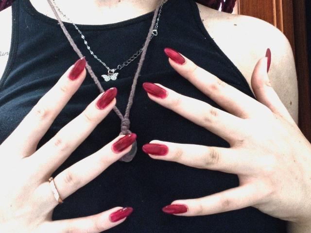 Le mani di Zanoza, con le unghie rosse e lucide, coprono i suoi seni, mentre indossa una canottiera nera e una collana d'argento con un ciondolo a forma di farfalla. Ha un sottile anello d'oro..