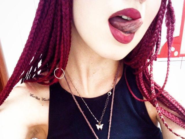 Zanoza, com cabelo trançado vermelho, língua de fora, batom vermelho escuro, blusa preta sem mangas, colar de ouro, tatuagem no ombro direito, brinco na orelha esquerda..