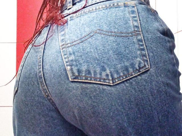 As nádegas de Zanoza, em calças jeans azuis justas com um bolso traseiro visível, exibindo suas curvas e formato arredondado. Tranças longas e vermelhas caem no lado esquerdo..