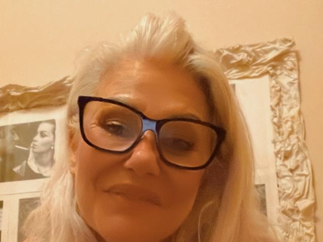 CrazyBionda with gray hair, wearing black glasses, smiling at the camera, has a cigarette in her mouth, picture of another person in the background.
