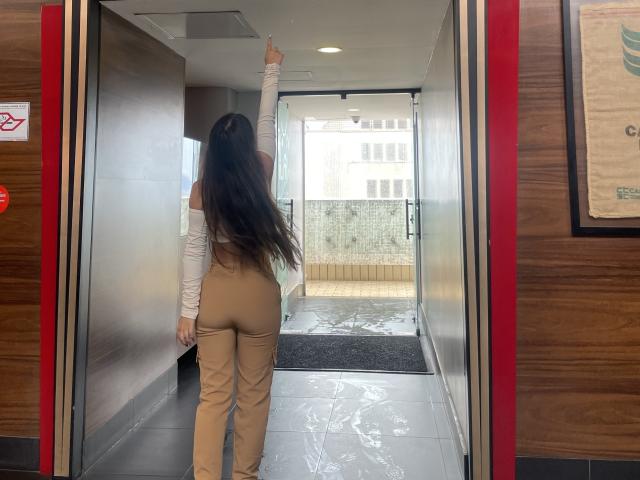 MeganWisse, con lunghi capelli castani, indossa una maglietta bianca con una spalla scoperta, pantaloni color carne, alza il braccio destro, mostrando i glutei, è in piedi nell'ingresso, con la schiena rivolta verso la telecamera, pareti di legno, porta di vetro, ambiente esterno luminoso..