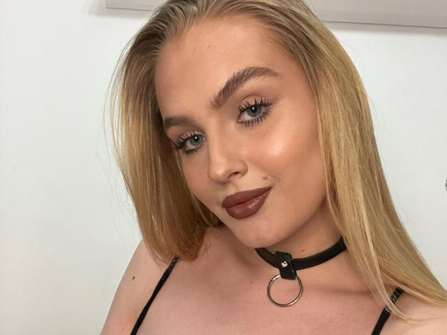 OliviaBent heeft blond haar, blauwe ogen en draagt donkere lipstick met een zwarte choker met een metalen ring. Haar borsten zijn klein en ze kijkt naar de camera..