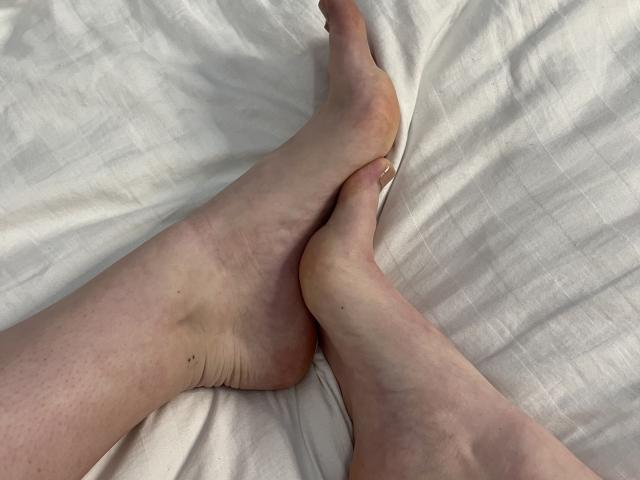 OliviaBent's bleke voeten zijn gekruist op een wit bed, waarbij licht gerimpelde zolen en tenen met een rozeachtige huid zichtbaar zijn. Haar nagels zijn kort en schoon..
